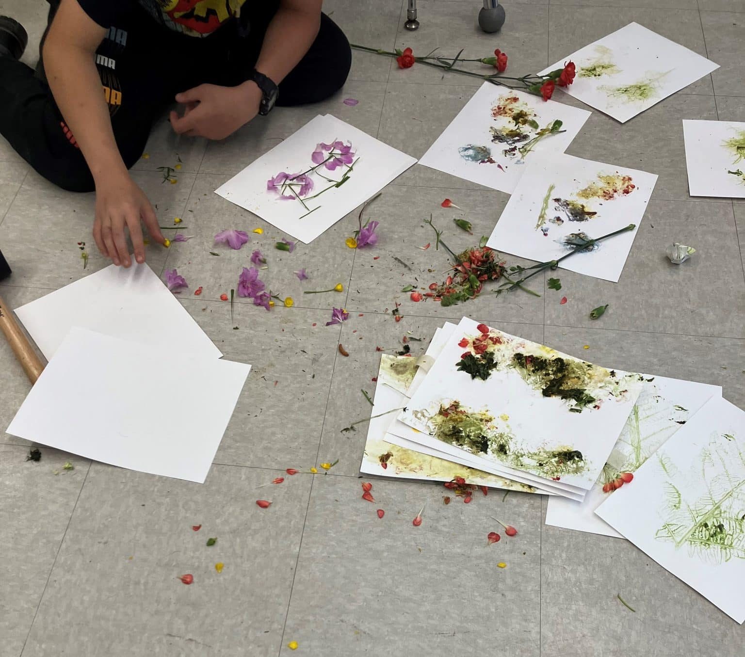 Unidentified student creates a new piece of 'plant pounding' artwork, extracting plant pigments by hammering to make prints, surrounded by previous creations.
