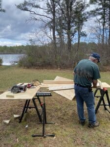 An individual working with wood, using tools outdoors near a lake.